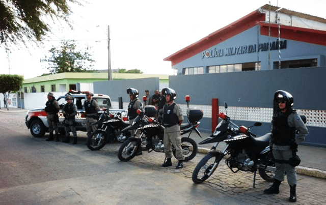 Caçada com três motos da Polícia Militar, cada uma com seu motorista uniformizado em preto, com a sede da polícia ao fundo